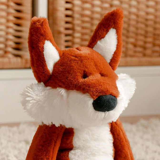 A plush fox toy sitting on a white shag rug in front of a wicker basket background. The toy has rust-orange fur, a white chest and inner ears, and a black nose.